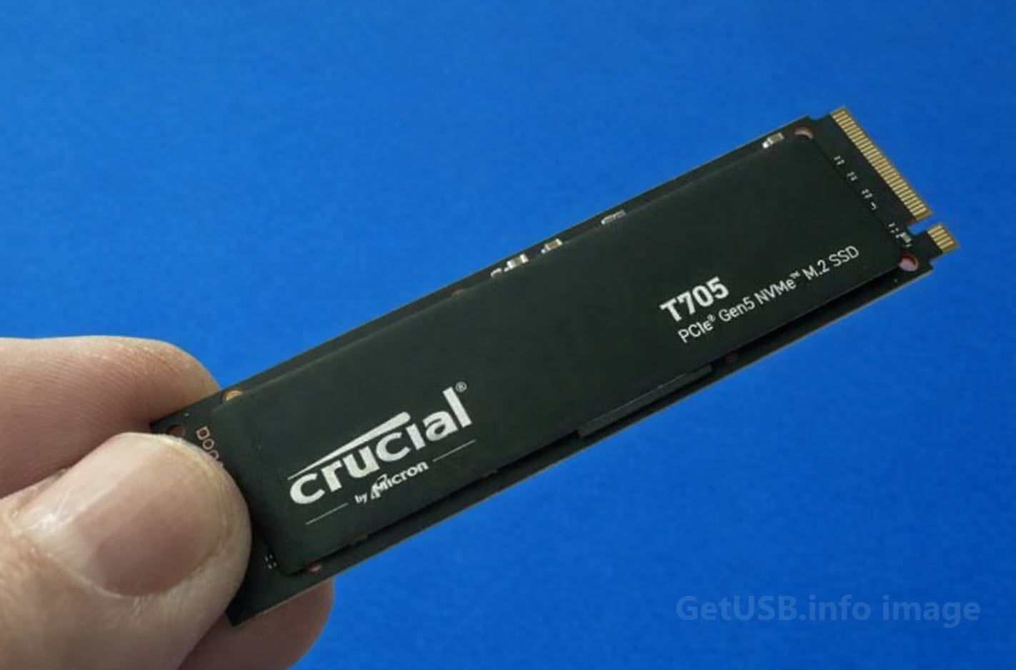 Close-up of a Crucial T705 PCIe Gen5 NVMe M.2 SSD held between fingers against a blue background